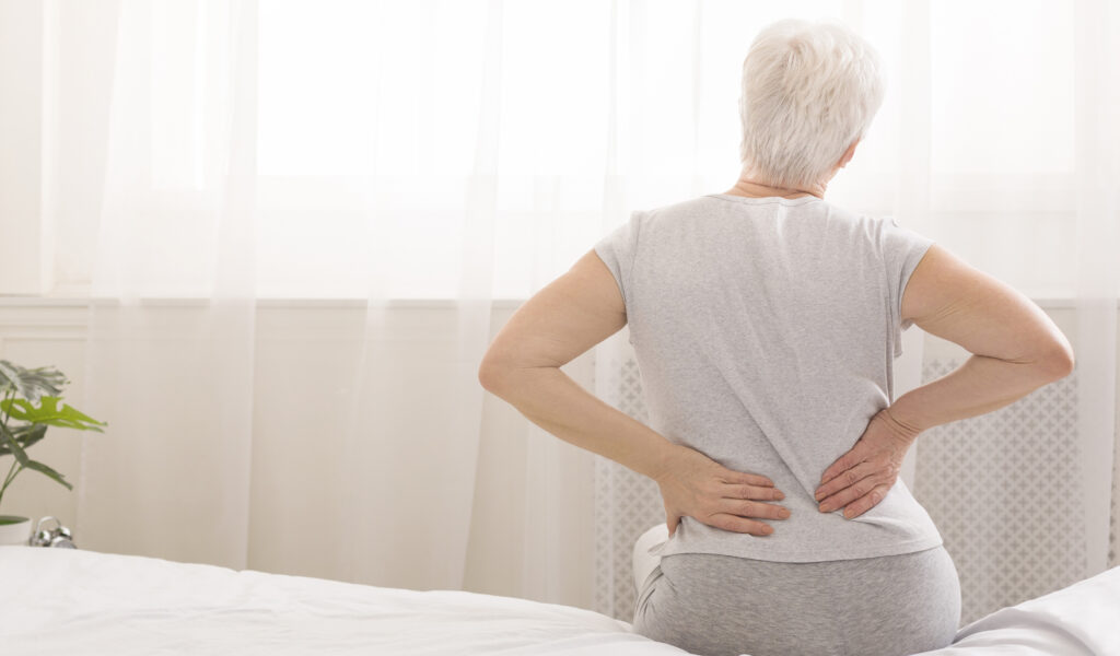 Senior woman sitting on the edge of a bed holding her lower back in pain